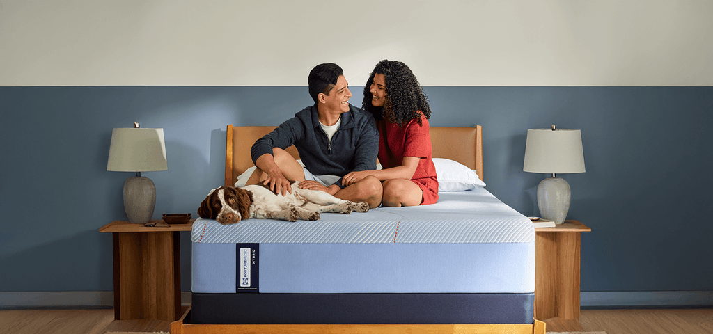 A man and woman sitting on a Posturepedic Hybrid mattress with their dog